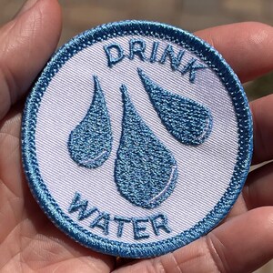 Könnte beinhalten: Ein weißer runder Patch mit blauer gestickter Kante. Der Patch zeigt drei blaue Wassertropfen und den Text "DRINK WATER" in blauer Stickerei.