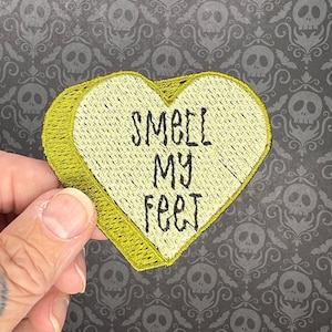 Op de afbeelding: Een gele hartvormige patch met de tekst "Smell My Feet" geborduurd in zwarte draad.