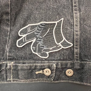 May include: Close-up of a black denim jacket with a hand patch. The patch is an embroidered design of a hand with a white outline and a black, textured center. The jacket has silver buttons and visible stitching.