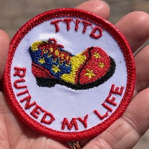 Puede incluir: Un parche blanco bordado con costuras rojas. El parche presenta un zapato de payaso rojo, amarillo y azul con el texto "TTID RUINED MY LIFE" alrededor del zapato.