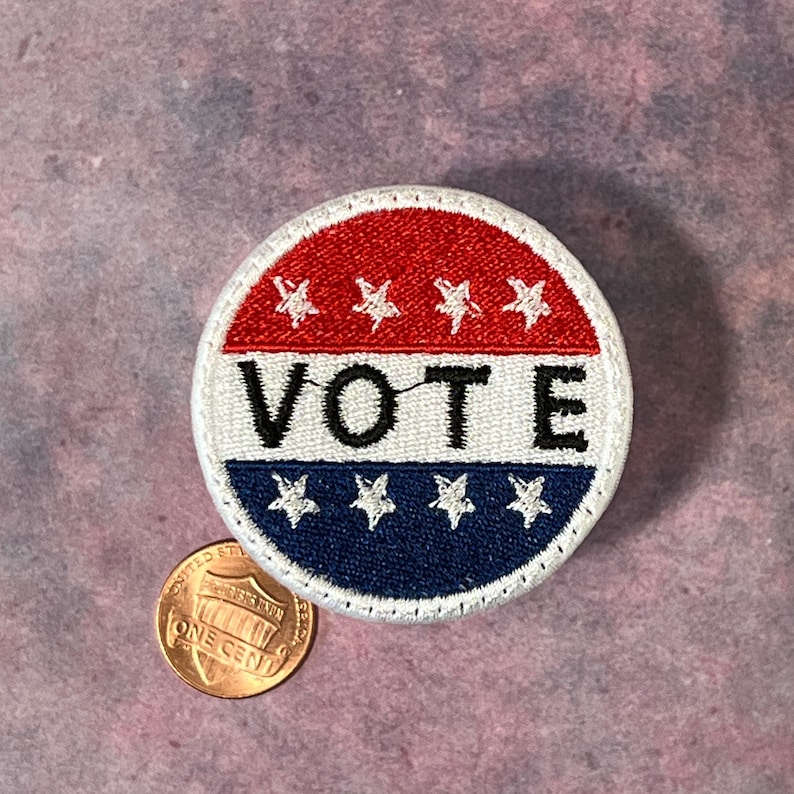 Peut inclure: Un &eacute;cusson rond avec le mot "VOTE" en lettres noires. La partie sup&eacute;rieure est rouge avec des &eacute;toiles blanches, le milieu est blanc et le bas est bleu avec des &eacute;toiles blanches. Une pi&egrave;ce de monnaie est &agrave; c&ocirc;t&eacute; de l'&eacute;cusson.