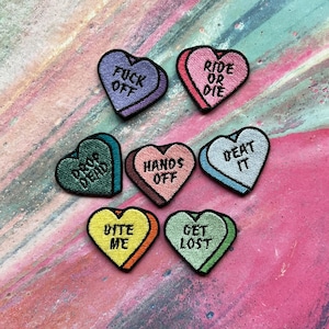 Może przedstawiać: Osiem haftowanych naszywek w kształcie cukierkowych serc z różnymi powiedzeniami, w tym "Drop Dead", "Fuck Off", "Ride or Die", "Hands Off", "Beat It", "Bite Me", "Get Lost".