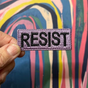 Può includere: Una toppa rettangolare lavanda con la scritta "RESIST" ricamata in filo nero. La toppa ha un bordo cucito. Lo sfondo presenta un design astratto con striature blu, rosa e gialle. Ideale per giacche o borse.