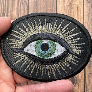 Peut inclure: Un patch brodé noir avec un œil vert avec une pupille bleue et un motif de soleil doré.