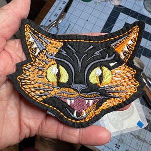 May include: Embroidered black cat patch with orange and yellow details. The cat has large yellow eyes, a snarling mouth with white teeth, and orange fur accents. The patch is outlined with a yellow stitch.