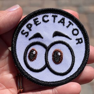 Könnte beinhalten: Ein runder, weißer Aufnäher mit dem Wort "SPECTATOR" in Schwarz, das sich oben wölbt. Darunter befinden sich zwei große Cartoon-Augen mit braunen Iris und schwarzen Umrissen sowie zwei schwarze Augenbrauen. Der Aufnäher hat einen schwarzen Rand.