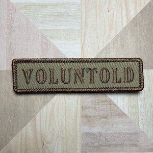 May include: A brown and green embroidered patch with the word "VOLUNTOLD" in brown thread.