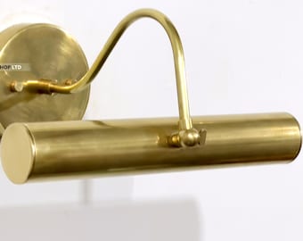 May include: A brass picture light fixture with a circular wall mount and a curved arm. The light has a cylindrical shade and is designed to illuminate artwork. The fixture is made of brass and has a warm, golden finish.