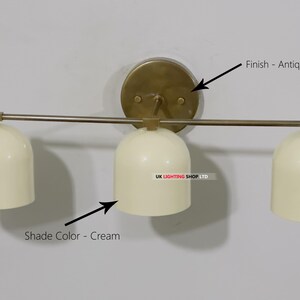 May include: Cream-colored, three-light wall sconce with an antique brass finish. The sconce features three bell-shaped shades and a circular backplate. The fixture is mounted on a light-colored wall, providing a clean and modern aesthetic.