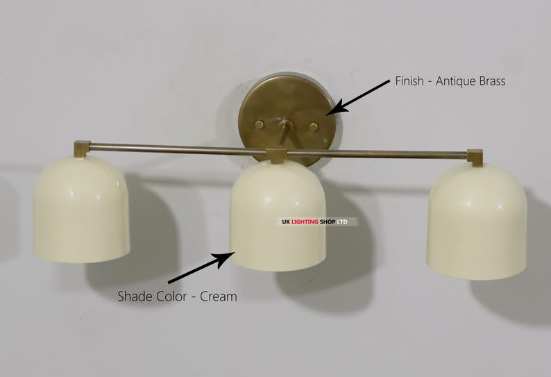 May include: Cream-colored, three-light wall sconce with an antique brass finish. The sconce features three bell-shaped shades and a circular backplate. The fixture is mounted on a light-colored wall, providing a clean and modern aesthetic.