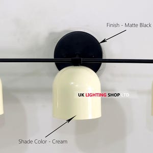 May include: A matte black wall-mounted light fixture with three cream-colored dome shades. The fixture has a circular base and a horizontal bar extending to hold the shades. The text "UK LIGHTING SHOP LTD" is visible.