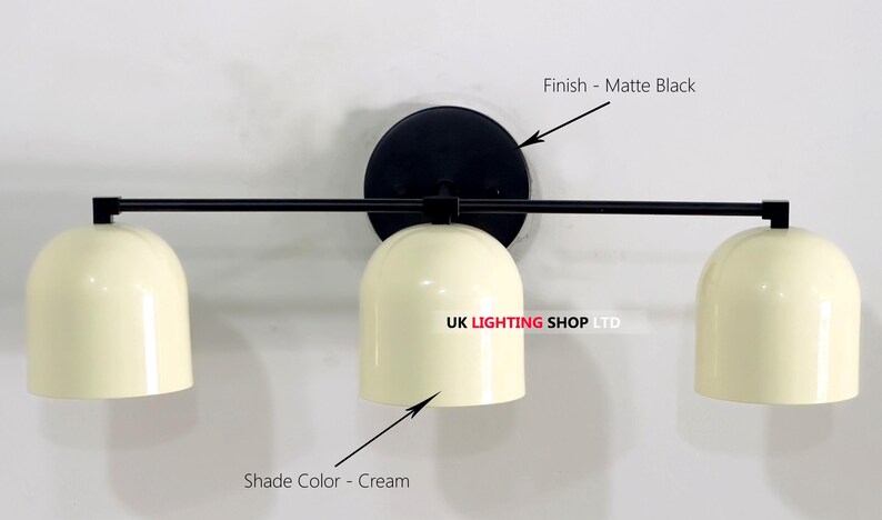 May include: A matte black wall-mounted light fixture with three cream-colored dome shades. The fixture has a circular base and a horizontal bar extending to hold the shades. The text "UK LIGHTING SHOP LTD" is visible.