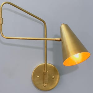 May include: A gold-toned wall sconce with a cone-shaped shade and an adjustable arm. The sconce has a circular base and is mounted to a wall.