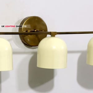 May include: A three-light wall sconce with a brass finish. The sconce features three cream-colored, dome-shaped shades. The fixture is mounted on a round brass backplate and has a horizontal bar.