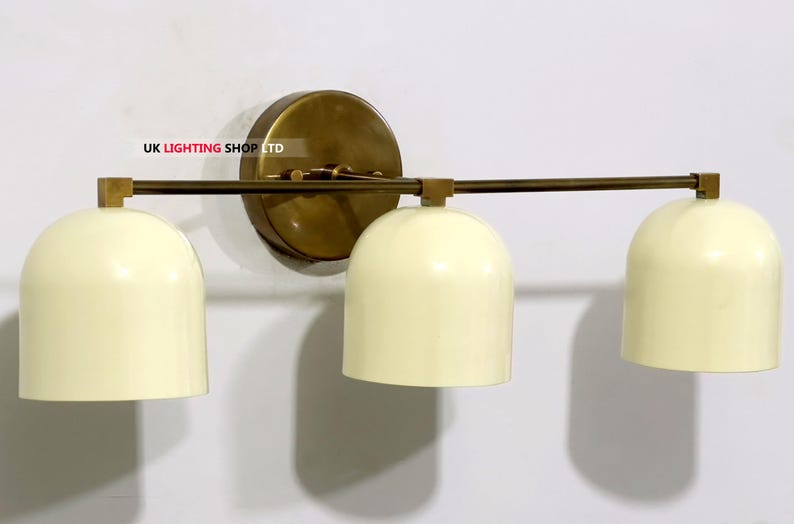 May include: A three-light wall sconce with a brass finish. The sconce features three cream-colored, dome-shaped shades. The fixture is mounted on a round brass backplate and has a horizontal bar.