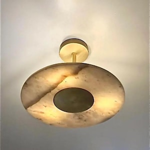 May include: A flush mount ceiling light fitting featuring a circular, translucent stone shade with natural veining in warm beige and brown tones. The centre of the shade has a circular brass plate, and it is attached to the ceiling by a brass canopy.