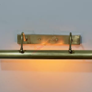 Puede incluir: Una lámpara de latón para cuadros con una placa trasera rectangular y una barra de luz cilíndrica. Dos brazos curvos conectan la barra de luz a la placa trasera. La luz está encendida, emitiendo un brillo cálido.