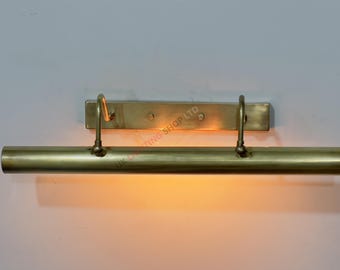 May include: A brass picture light fixture with a rectangular backplate and a cylindrical light bar. Two curved arms connect the light bar to the backplate. The light is on, casting a warm glow. The fixture is mounted on a light-colored wall.