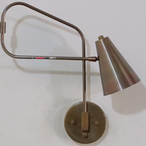 May include: A wall-mounted, adjustable lamp with a conical shade and a circular base. The lamp is made of metal with a bronze finish. The arm extends from the base and curves to hold the shade. The text "UK SHOP LTD" is visible on the arm.