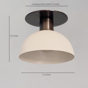 Puede incluir: Una lámpara de techo blanca en forma de cúpula con una cubierta de metal negro y un acabado bronce. La lámpara tiene un diámetro de 15 cm y una altura de 15 cm.