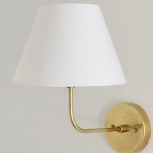 May include: A wall-mounted lamp featuring a white lampshade and a brass-toned metal arm and base. The lampshade has a conical shape. The base is circular and fixed to the wall. The arm curves to hold the lampshade.