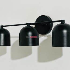 May include: A black, three-light wall sconce with a minimalist design. The fixture features three bell-shaped shades, a horizontal bar, and a circular base, all in a matte black finish. The sconce is mounted on a white wall.