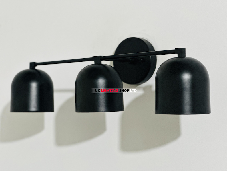 May include: A black, three-light wall sconce with a minimalist design. The fixture features three bell-shaped shades, a horizontal bar, and a circular base, all in a matte black finish. The sconce is mounted on a white wall.