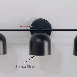 May include: A matte black three-light wall sconce. The fixture has a circular base, a horizontal bar, and three bell-shaped shades. The text "Full Matte Black" is below the center shade. The sconce is mounted on a white wall.