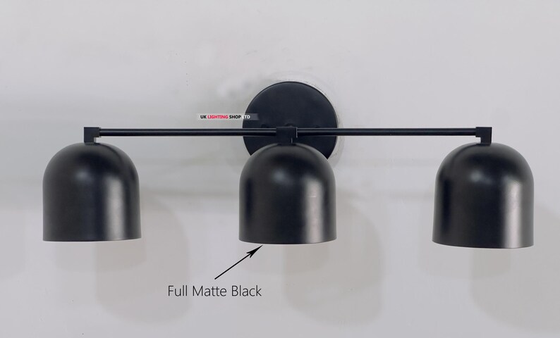May include: A matte black three-light wall sconce. The fixture has a circular base, a horizontal bar, and three bell-shaped shades. The text "Full Matte Black" is below the center shade. The sconce is mounted on a white wall.