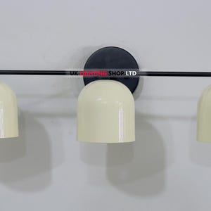 May include: A modern wall-mounted light fixture with three cream-colored, dome-shaped shades. The shades are attached to a black horizontal bar, which is connected to a circular black base. The fixture is mounted on a white wall.