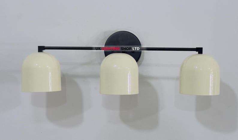 May include: A modern wall-mounted light fixture with three cream-colored, dome-shaped shades. The shades are attached to a black horizontal bar, which is connected to a circular black base. The fixture is mounted on a white wall.