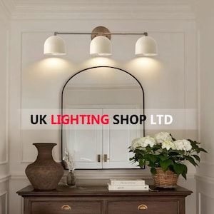 May include: A three-light vanity fixture with white, bell-shaped shades and a brushed nickel finish. The fixture is mounted above a black-framed mirror. The text "UK LIGHTING SHOP LTD" is displayed.