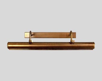 May include: A horizontal brass picture light fixture featuring a long cylindrical tube attached to a rectangular wall-mounting plate by two curved adjustable arms. The metal surface has a polished finish with a warm golden metallic tone.