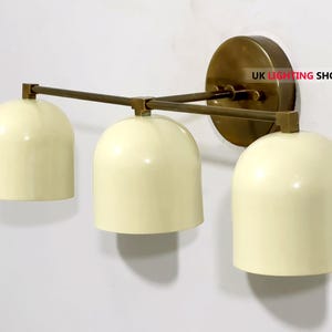 May include: Cream-colored, three-light wall sconce with a brass finish. The sconce features three bell-shaped shades and a circular backplate. The arms and backplate are a warm brass color, and the shades are a soft cream color.
