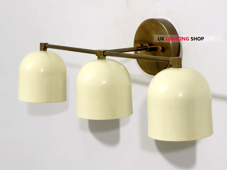 May include: Cream-colored, three-light wall sconce with a brass finish. The sconce features three bell-shaped shades and a circular backplate. The arms and backplate are a warm brass color, and the shades are a soft cream color.