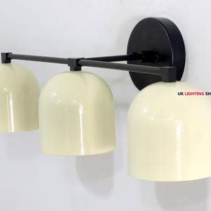 May include: A modern wall-mounted light fixture with three cream-colored dome shades. The fixture has a black mounting plate and a black horizontal bar connecting the shades. The text "UK LIGHTING SHOP LTD" is visible.