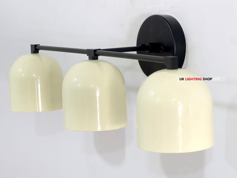 May include: A modern wall-mounted light fixture with three cream-colored dome shades. The fixture has a black mounting plate and a black horizontal bar connecting the shades. The text "UK LIGHTING SHOP LTD" is visible.