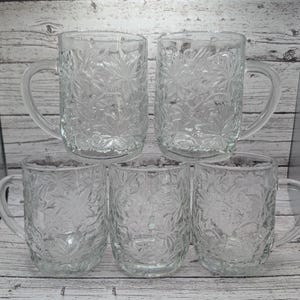 May include: Five clear glass mugs with a leaf pattern are stacked on a wooden surface. Each mug has a handle and a cylindrical shape. The mugs are made of clear glass, allowing light to pass through. The leaf pattern adds a decorative touch to the glassware.
