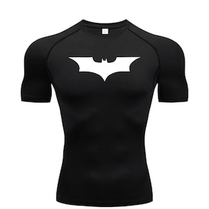 May include: Black compression shirt featuring a white Batman logo on the chest. The short-sleeved shirt has a crew neck and is designed for athletic wear. The logo is a stylized bat symbol.