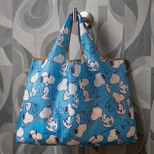 May include: A blue reusable shopping bag featuring a repeating pattern of Snoopy characters in various poses. The bag has brown trim and handles, and is hanging from a silver hook against a patterned gray background.