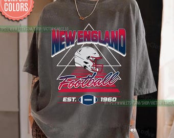 May include: A gray t-shirt featuring a New England Football design. The design includes the words "New England" in blue and red, a football helmet, and the word "Football" in red script. The shirt also has "EST. 1960" and a football graphic.