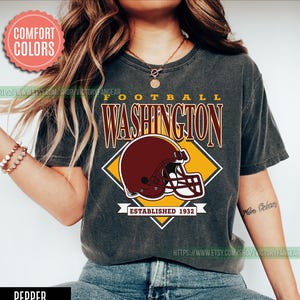 May include: Pepper-colored t-shirt with a football-themed graphic. The design features the word "FOOTBALL WASHINGTON" in maroon lettering, a maroon helmet, and the text "ESTABLISHED 1932". The shirt has a vintage, worn look.