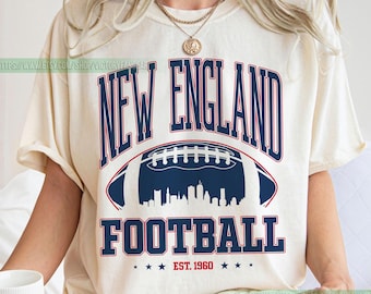 May include: Ivory-colored t-shirt featuring the text "NEW ENGLAND FOOTBALL" in navy blue, with a football graphic and a skyline silhouette. The shirt includes the text "EST. 1960" and stars. The shirt is a short-sleeved, crew neck style.