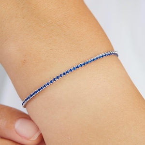 May include: A delicate silver bracelet with a row of small, blue gemstones. The bracelet is shown on a person's wrist.