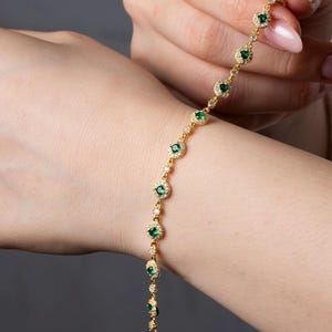 Op de afbeelding: Een gouden armband met een ketting en een rij groene smaragdstenen omringd door kleinere heldere stenen.