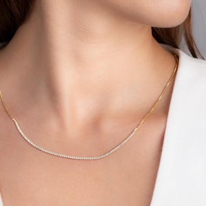 May include: A delicate gold necklace with a curved bar of small, sparkling gemstones. The necklace is worn around the neck, showcasing the subtle elegance of the jewelry. The gemstones are set closely together, creating a continuous line of brilliance.