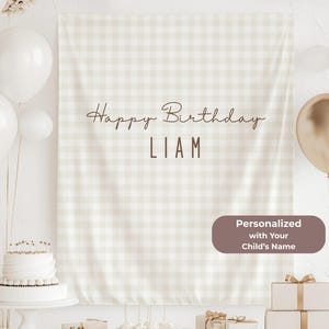 May include: A beige and white checkered backdrop with the text "Happy Birthday LIAM" in brown script. The backdrop is decorated with white and gold balloons, a cake, and gifts. The text "Personalized with Your Child's Name" is also present.