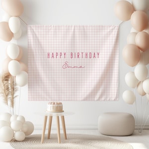 May include: A birthday party scene with a pink and white checkered backdrop that reads "HAPPY BIRTHDAY Emma". The scene includes white and blush-colored balloons, a small cake on a white table, and a neutral-colored ottoman.