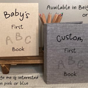 May include: Two fabric-covered photo albums, one beige and one gray, with the text "Baby's First ABC Book" on the beige album and "Custom First ABC Book" on the gray album. The text "Available in Beige or Grey" is written above the albums.