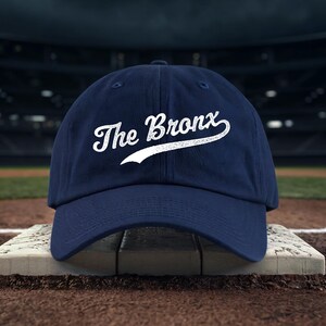 Puede incluir: Gorra de béisbol azul marino con la inscripción "The Bronx" en blanco. La gorra está colocada en un campo de béisbol. Un accesorio clásico e informal, para deportes o uso diario.